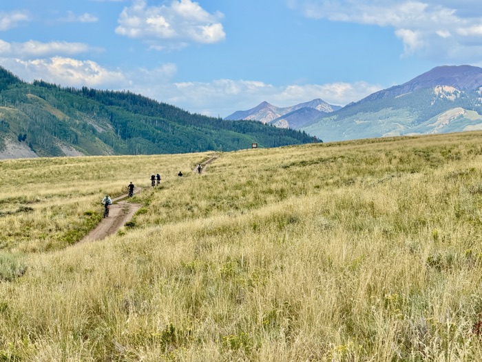 Riders strung out along singletrack crossing an open meadow below the Elk Mountains
